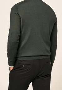 Ciemnozielony sweter z dzianiny z fakturowanymi łatami na łokciach, zestawiony z czarnymi spodniami posiadającymi tylne kieszenie i ozdobne guziki.