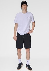 Weißes T-Shirt mit kleinem rotem Logo, schwarze Shorts, schwarze Sneakers und weiße Sportsocken. Legeres Outfit mit lockerem Sitz und elastischem Bund.