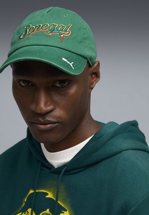 Man wearing a green baseball cap with "Senegal" embroidered in yellow, orange, and green, paired with a green hoodie featuring a yellow graphic.