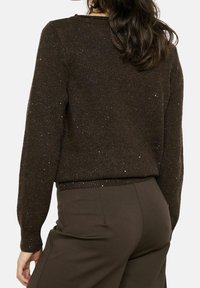 Vue de dos d'une femme portant un pull en tricot marron foncé avec des taches, assorti à un pantalon marron foncé, sur un fond uni.