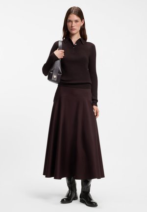 Femme debout portant une longue jupe marron foncé, un pull polo à manches longues assorti, des bottes noires et tenant un sac à épaule noir.