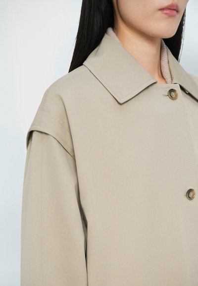 Μπεζ παλτό με μεγάλο γιακά, κούμπωμα με κουμπιά και ορατές λεπτομέρειες ραφής. Λείος υφασμάτινος χαρακτήρας με άνετη εφαρμογή.