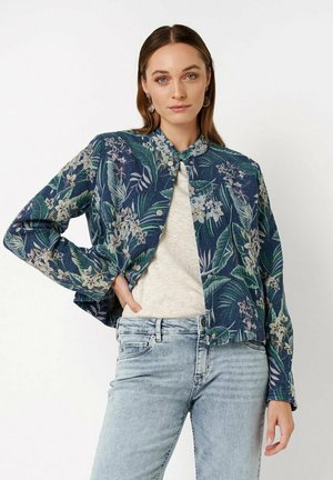 Frau trägt eine dunkelblaue, florale Knopfjacke über einem hellen Hemd mit hellen Jeans, posiert mit einer Hand in der Hüfte.