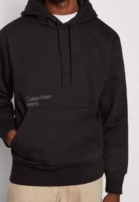 Man i svart Calvin Klein Jeans hoodie med frontficka och dragsko, i kombination med beiga byxor.