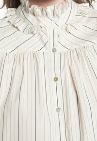 Krämfärgad blus med tunna, vertikala ränder i beige och marinblå. Ryllad krage, knappfram med cirkulära grönaktiga knappar och veckade detaljer.