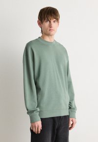 Sudadera verde claro con cuello redondo y puños acanalados. Presenta un pequeño logo sutil en el pecho. Combinada con pantalones oscuros.