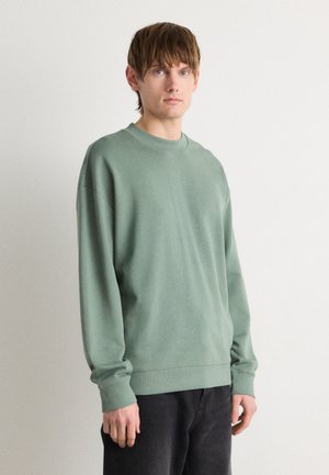 Sweatshirt vert clair avec un col rond et des poignets côtelés. Présente un petit logo discret sur la poitrine. Porté avec un pantalon foncé.
