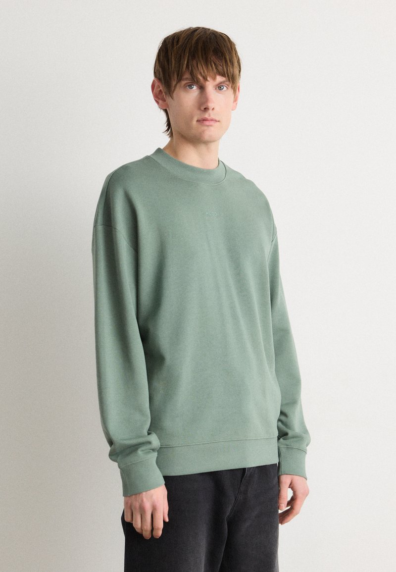 Sudadera verde claro con cuello redondo y puños acanalados. Presenta un pequeño logo sutil en el pecho. Combinada con pantalones oscuros.