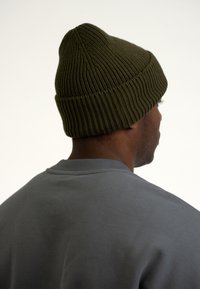 Căciulă tricotată verde cu textură ribbed, vârf conic, purtată de o persoană cu un hanorac gri, arătând spatele capului și gâtul.
