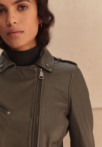 Giacca in pelle verde oliva con chiusura a zip, bottoni a pressione e tasca con cerniera sul lato sinistro. Colletto alto e spalle strutturate.