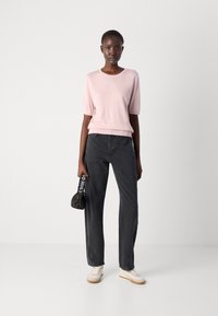Lys rosa kortermet genser, avslappet passform; kombinert med høyt liv, mørkegrå vide jeans; holder en svart veske med hvit tekst.