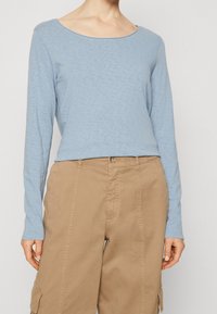 Světle modrá dlouhá košile s vykrojeným výstřihem, vyrobená z texturované látky. V kombinaci s khaki volnými cargo kalhotami.