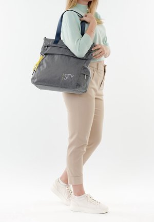 Woman wearing light green sweater and beige pants holding large gray tote bag with black straps and yellow zipper pull.