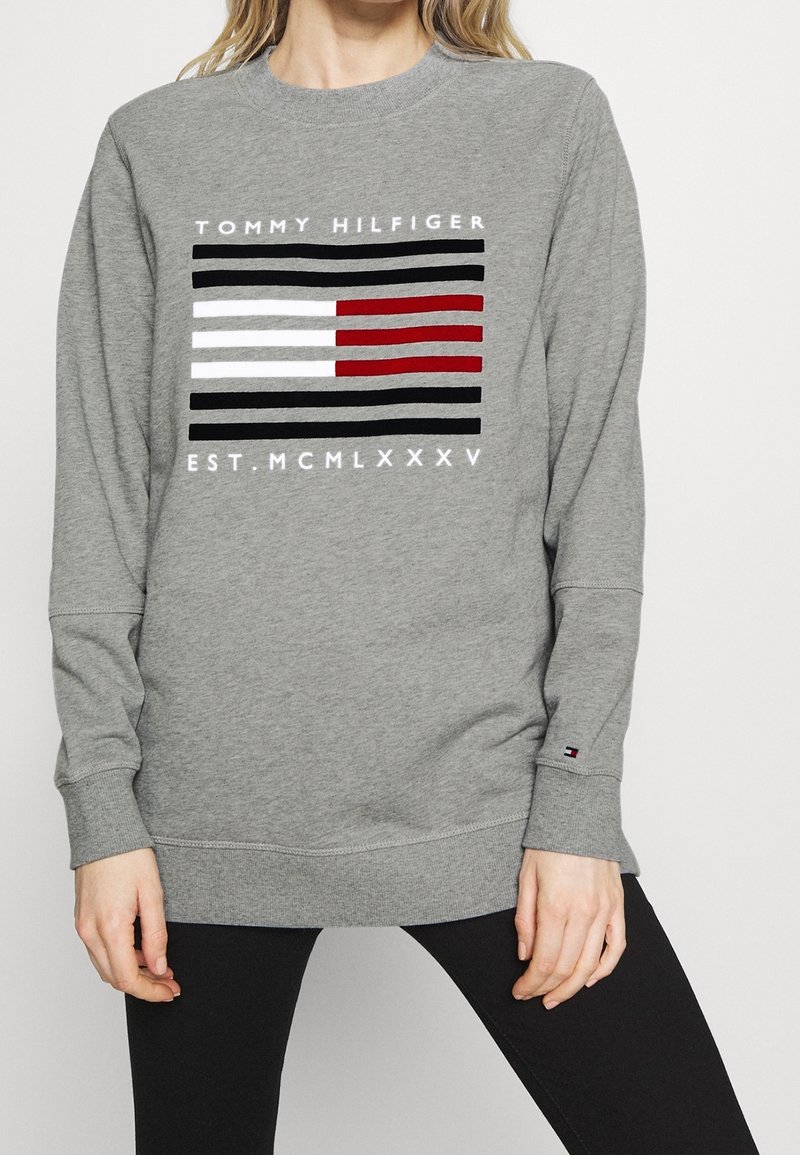 Personne portant un sweat-shirt gris avec le logo Tommy Hilfiger comportant des bandes horizontales noires, blanches et rouges, et le texte "EST. MCMLXXXV" en dessous.