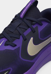 Κοντινή λήψη μοβ και μαύρου παπουτσιού Nike για τρέξιμο με υφασμάτινο δίχτυ με υφή, κορδόνια και μεγάλο μπεζ λογότυπο Swoosh στο πλάι.