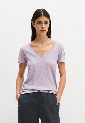 Woman with long dark hair wearing a light purple V-neck T-shirt and dark drawstring pants, standing with hands in pockets against a plain background.