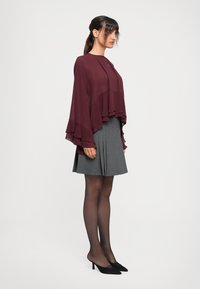 Blusa leggera bordeaux con maniche larghe e sovrapposte abbinata a una gonna grigia plissettata e collant scuri trasparenti. Tacchi a punta nera.