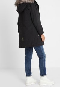 Sort parka-jakke med faux fur-beklædning, lynlås foran, sidelommer og justerbar hætte. Båret med blå jeans og hvide støvler.