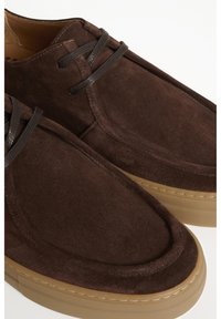 Smeđe suede cipele s okruglim vrhom, šivanjem detalja i tamnim konopcem za vezivanje. Imaju teksturiranu svijetlosmeđu gumenu potplat.