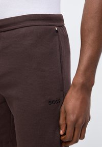 Κοντινό πλάνο ενός ατόμου που φοράει σκούρα καφέ σόρτς με υφή, με πλαϊνή τσέπη με φερμουάρ και λογότυπο "BOSS", το χέρι χαλαρό δίπλα στον μηρό.