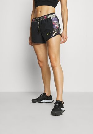 Athletische Frau trägt schwarze Sportshorts mit bunten Seitenstreifen, schwarzen Sport-BH und schwarze Laufschuhe und steht vor einem schlichten Hintergrund.
