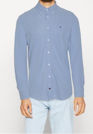 Mann trägt ein hellblaues, langärmliges Hemd mit kleinem Tommy Hilfiger Logo und hellblaue Jeans.