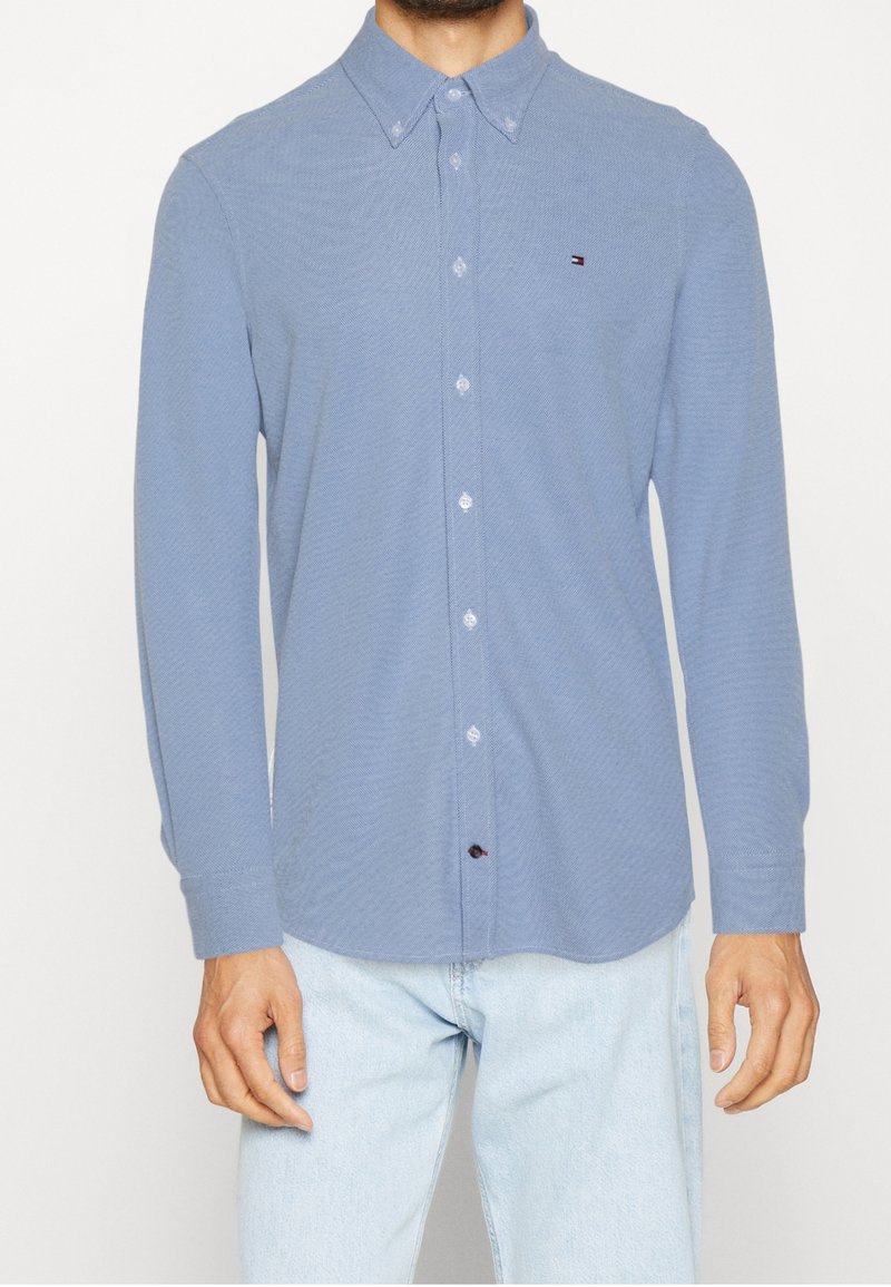 Homme portant une chemise à manches longues bleu clair avec un petit logo Tommy Hilfiger et un jean bleu clair.