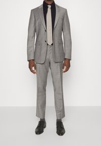 Šedý oblek s přizpůsobeným střihem, dvěma knoflíky a klopami s výřezem, doplněný černou košilí a světle šedou kravatou. Černé společenské boty dokončují vzhled.