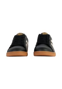 Zapatillas de cuero negro con acentos blancos, suela de goma marrón plana, logo prominente en la lengüeta, diseño con cordones, textura suave.