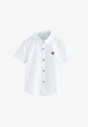 Niño pequeño con camisa blanca de manga corta con parche de animal, pantalones cortos beige y zapatos beige sin cordones, mirando hacia abajo.