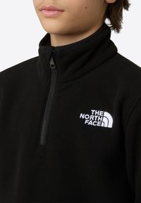 Schwarzer Fleece-Pullover mit hohem Kragen und halbem Reißverschluss. Mit weißem, besticktem Logo auf der linken Brust. Weiche Textur.