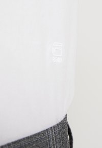 Biała bawełniana tkanina o subtelnej teksturze, z małym haftowanym logo. Szary wzór w kratę na dole, podkreślający wizualny kontrast.