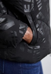 Zwarte pufferjack met subtiele tonale geometrische patronen, voorzien van een ritszak en een gladde, matte textuur.
