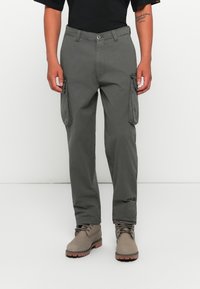 Man in een donkergrijze cargobroek en taupekleurige veterschoenen, staand tegen een witte achtergrond met ontspannen handen langs de zij.