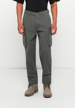 Man wearing dark gray cargo pants and taupe lace-up boots, standing against a white background with hands relaxed by sides.