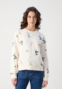 Lys beige sweatshirt med blomsterbroderi, som har grønne og gule planter. Klassisk rund hals og ribbestrikkede ermer. Denim jeans synlige.
