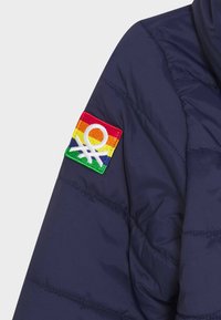 Veste matelassée navy avec une texture en quilt, ornée d'un patch brodé coloré en rouge, orange, jaune, vert et bleu sur la manche.