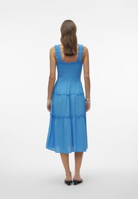 Robe bleue à volants avec un corsage smocké et des bretelles à volants, présentant une texture fluide et une longueur mi-mollet. Design simple, sans motifs.