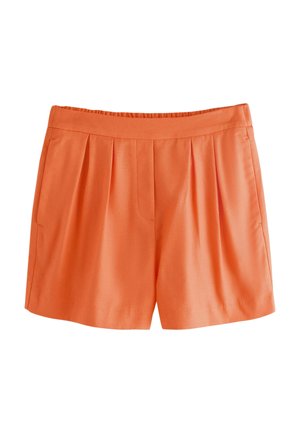 Hosszú barna hajú nő narancssárga ujjatlan mellényt és hozzáillő rövidnadrágot visel, bent áll egy nagy ablak mellett, amelyből kinti természet látványa tárul elénk.