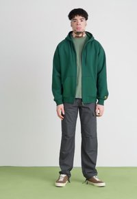 Grüne Zip-Hoodie über einem hellgrünen T-Shirt, kombiniert mit dunkelgrauen Cargo-Hosen und braunen Sneakers. Strukturierter Stoff, lässige Passform, verstellbare Kapuze.