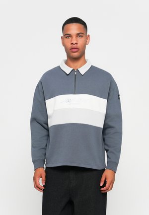 Jonge man in een grijs-witte sweatshirt met kwartrits en tekst op de borst, staand tegen een eenvoudige witte achtergrond.