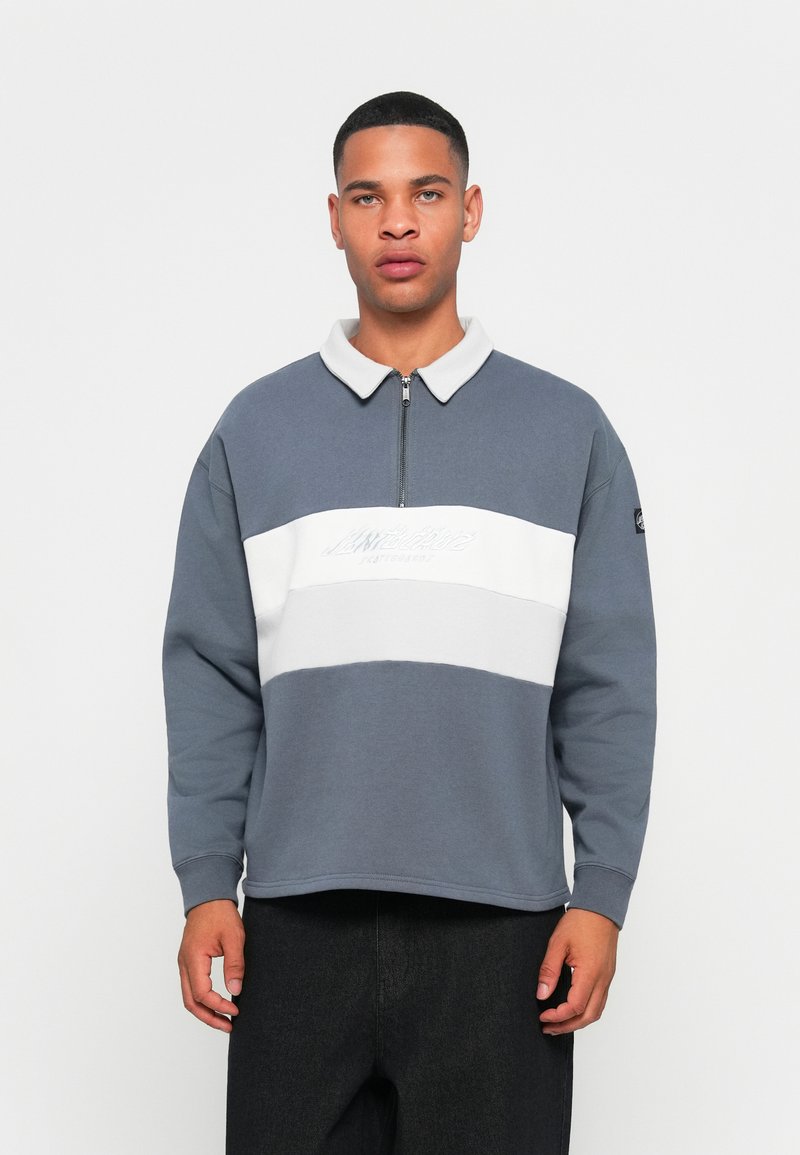 Ung mand iført en grå- og hvid halvzip-sweatshirt med tekst på brystet, stående foran en ensfarvet hvid baggrund.