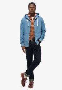 Felpa azzurro chiaro con cordini bianchi sopra una camicia a quadri arancione, abbinata a jeans scuri e scarpe di pelle marroni con dettagli grigi.