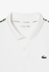 Biała koszulka polo wykonana z fakturowanej tkaniny, z zielonym wykończeniem kołnierzyka oraz małym zielonym logo krokodyla na piersi. Klasyczny krój.