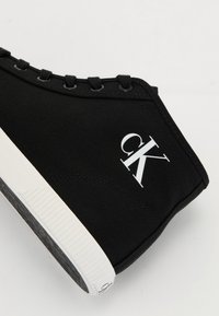 Svart canvas högsko med vit gummisula. Har "CK"-logotyp i vitt på sidan och svarta snören framtill.