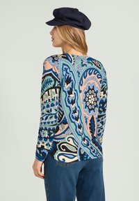 Haut à manches longues avec un imprimé cachemire multicolore en bleu, vert et rose. Le tissu a une texture lisse. Porté avec un jean bleu et une casquette marine.