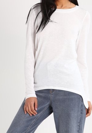Camicia bianca a maniche lunghe con scollo rotondo e orlo leggermente curvo, realizzata in tessuto leggero e semi-trasparente. Abbinata a jeans blu.