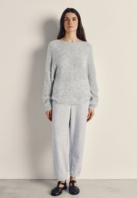 Žena stojaca opretá o béžovú stenu, oblečená v svetlosivom chlpatom svetri, zladených teplákoch a čiernych tkaných sandáloch.