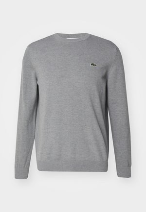 Grå strikkegenser med lange ermer, ribbestrikket rund hals og mansjetter, med en liten grønn Lacoste-logo på brystet. Myk tekstur.