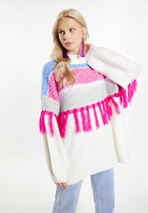 Blonde Frau trägt übergroßen weißen Pullover mit blauem und pinkem Muster und pinken Fransen, posiert mit Hand am Hals, hellblaue Jeans.